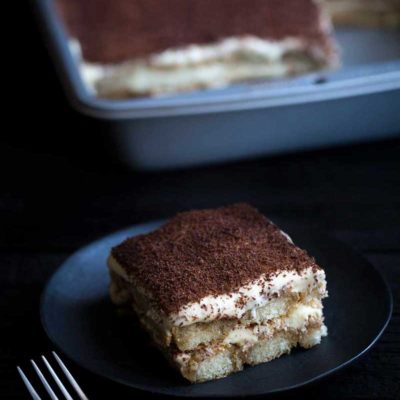 Closeup of tiramisu serving on a plate with full pan of tiramisu in the background