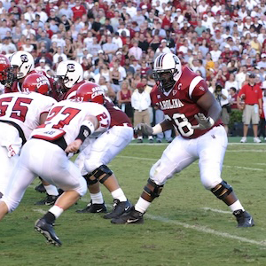 football player in white helmet, garnet jersey and white pants backpedals during a game