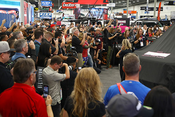 Group of people with cameras watching a presentation at a SEMA Show booth.