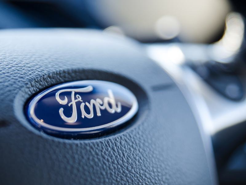 A close-up photograph showing a car steering wheel with the blue ‘Ford’ logo in the centre of it. The background is blurred.