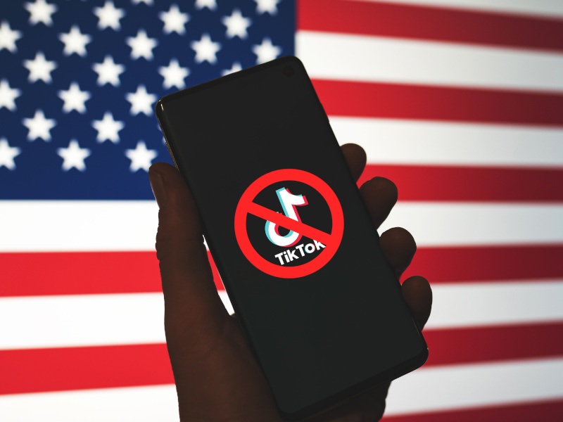 An American flag in the back with a hand holding a smartphone that shows a cross over the TikTok app logo.