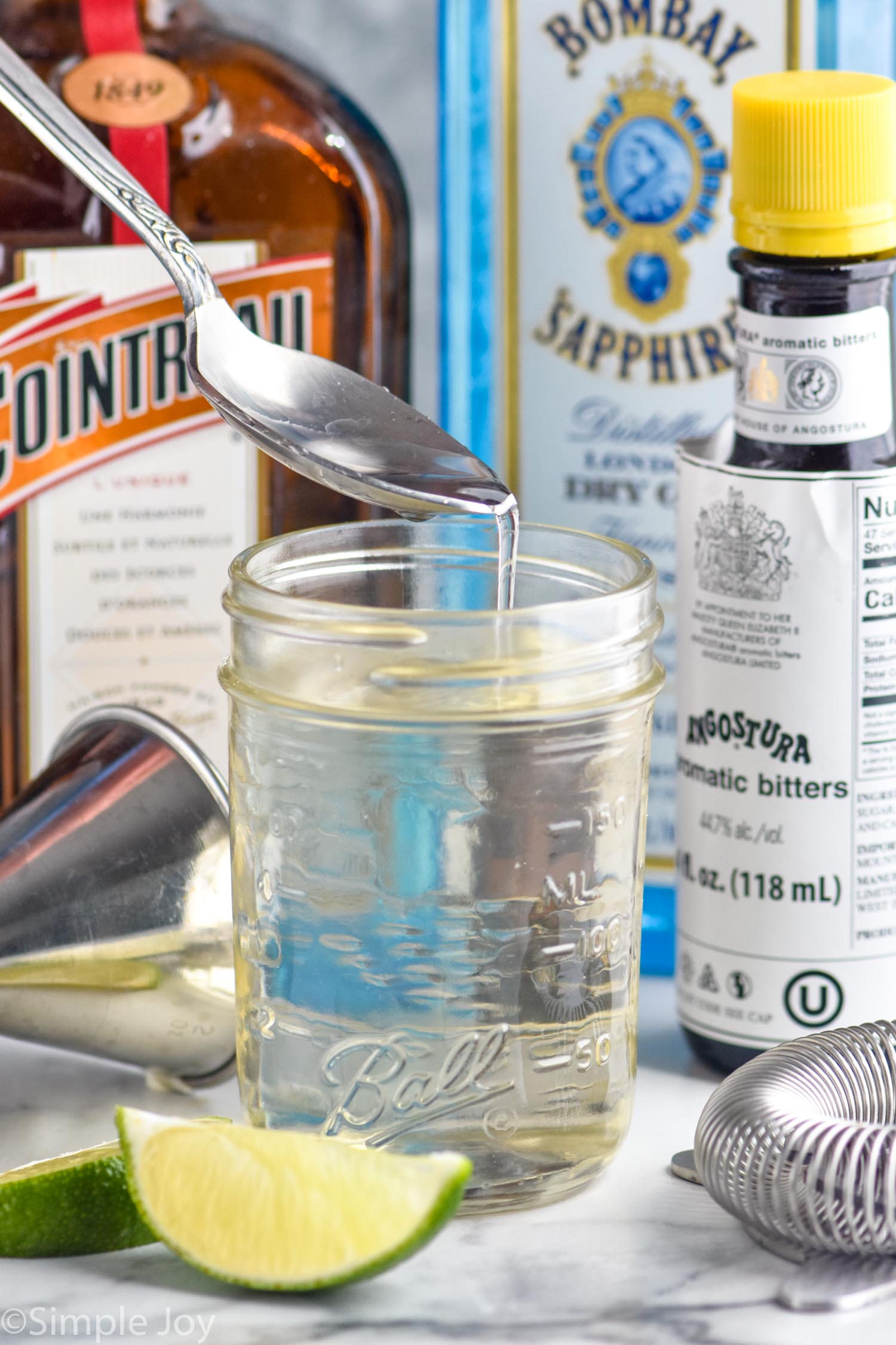 Photo of a spoon over a jar of Simple Syrup recipe. Lime wedges, cocktail jigger, and bottles of cointreau, gin, and bitters beside jar.