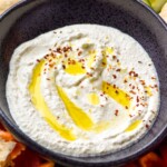 Bowl of Whipped Feta Dip topped with olive oil and red pepper flakes with cucumber slices, red pepper slices, and pita bread surrounding