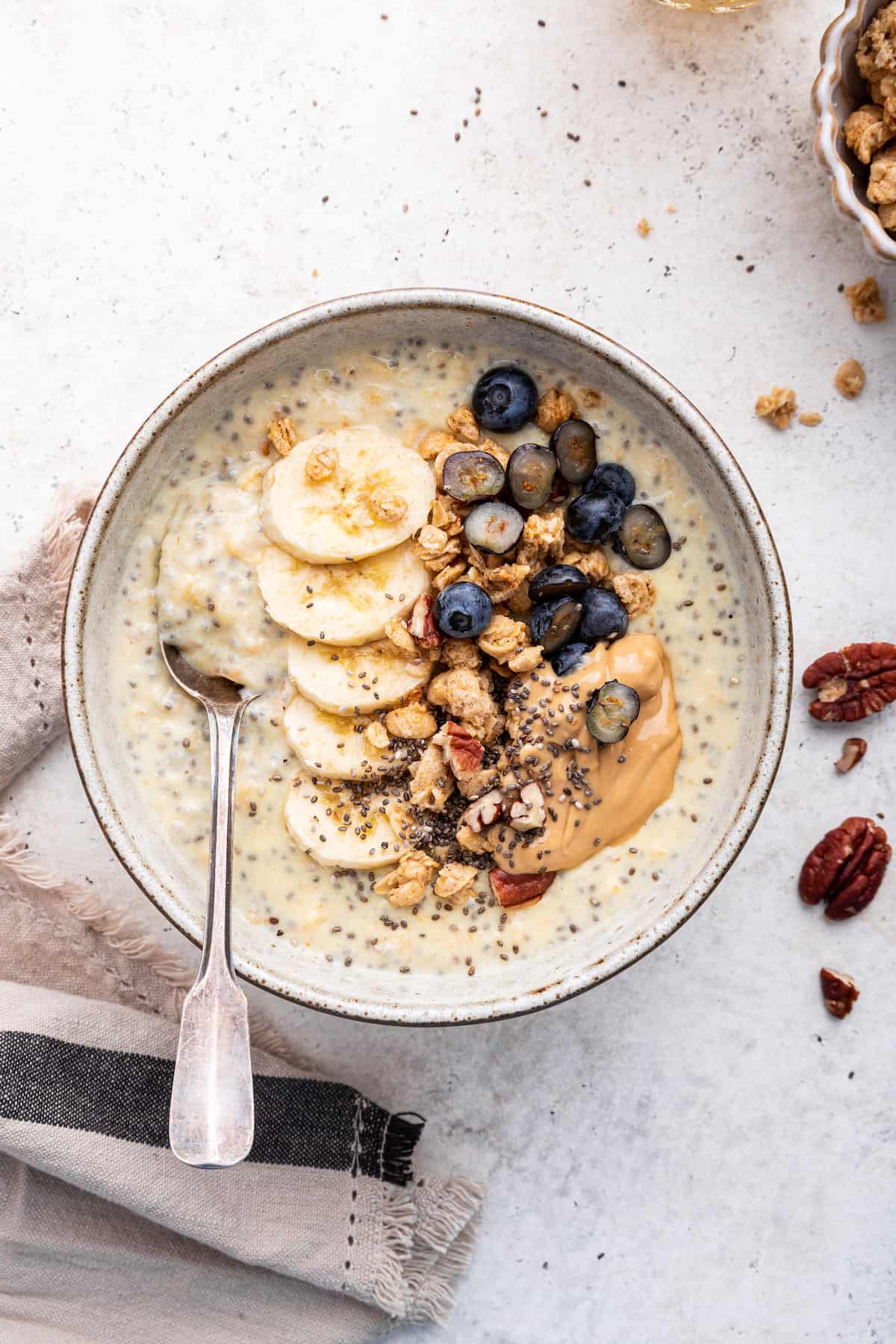 Overhead view of spoon in bowl of protein oatmeal topped with bananas, peanut butter, blueberries, pecans, and chia seeds