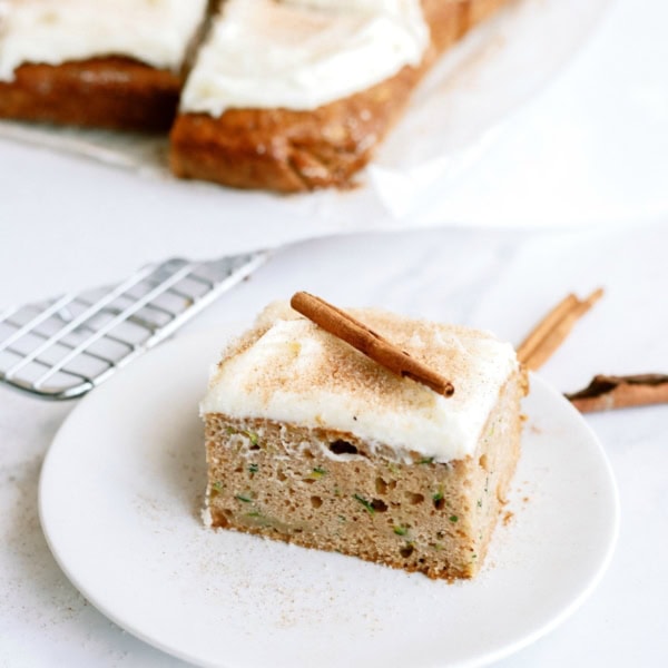 A slice of cake topped with frosting and a cinnamon stick is placed on a white plate, with the rest of the cake in the background.