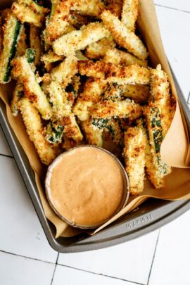 A lined sheet pan crispy zucchini fries and a small cup of dipping sauce.