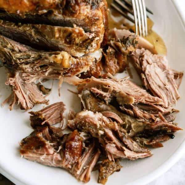 Cooked Oven Roasted Pork Shoulder on a white serving plate with two forks shredding some of the meat.