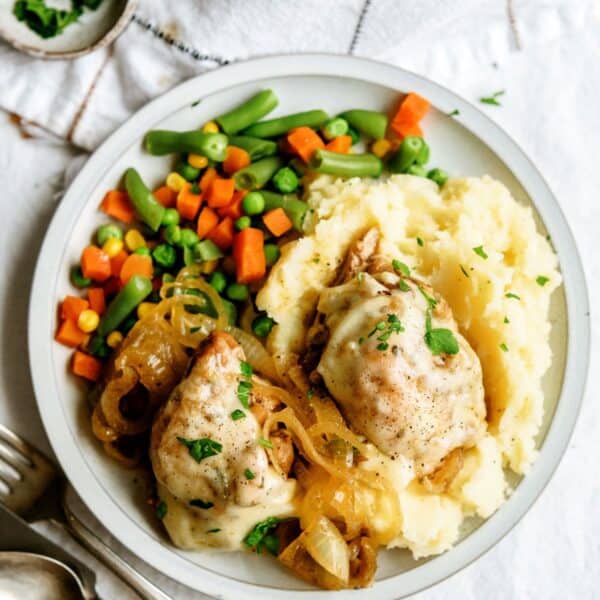 A plate containing mashed potatoes, two pieces of cooked chicken topped with onions, and a side of mixed vegetables including green beans, peas, carrots, and corn.