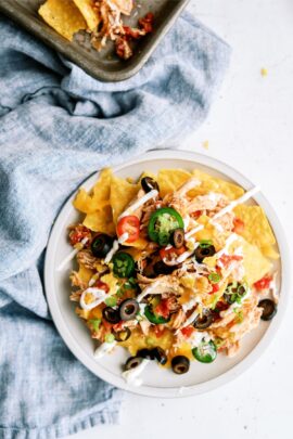 A plate of nachos topped with shredded chicken, sliced jalapeños, black olives, green onions, cherry tomatoes, and drizzled with white sauce, placed on a blue cloth.