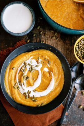 A bowl of Acorn Squash Soup with cream sauce and toasted pumpkin seeds on top. Separate bowls of Acorn Squash Soup, pumpkin seeds and cream sauce in the background.