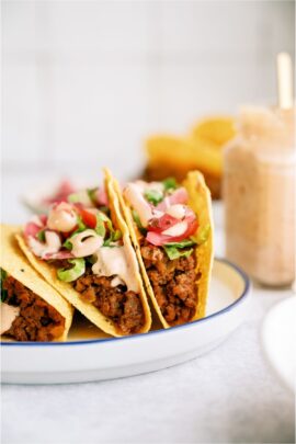 3 Cheeseburger Tacos on a plate with toppings and sauce. A jar of sauce with a spoon in it on the side.