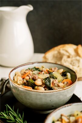 A bowl of Tuscan White Bean Soup with a white pitcher and bread on a plate in the background.