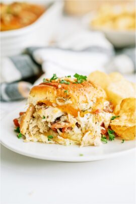 Crack Chicken Slider sandwich on a white plate with chips. Pan of Crack Chicken Sliders in the background.