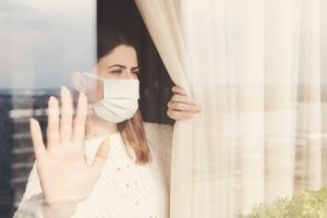 woman with face mask gazing out of a window with her hand pressed against the glass