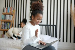 Two girls are sitting on a bed in a shared bedroom. One girl chats with someone on a computer. The other girl watches something in the background.