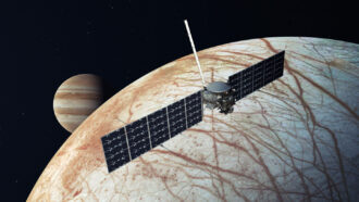 an illustration shows the Europa Clipper in front of Europa (one of Jupiter's moons), which is light-colored crisscrossed with brownish cracks. Jupiter is visible in the distance