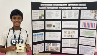 Samvith Mahadevan holds a small white device designed to detect food allergens and stands next to a poster board describing the project.