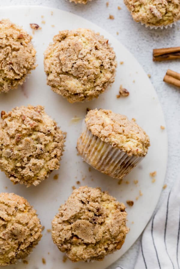 Six coffee cake muffins sit on a white serving platter with crumbs on the side.