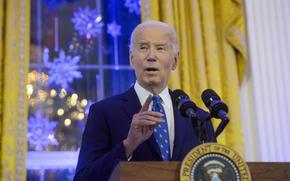 A man in a navy suit and blue tie speaks at a podium.