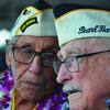 Two elderly men wearing hats and flowered leis speak to each other.