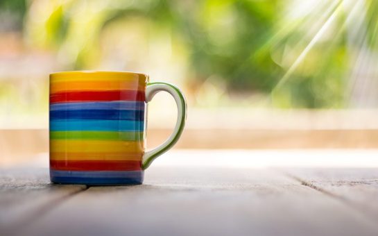 A mug of tea with the rainbow flag painted on it sits on a wooden table.