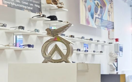 The image shows a gold statue placed on a pedestal inside an opticians. The statue features a winged design integrated with a triangular shape, symbolising an award. Behind the statue, shelves display a variety of eyeglasses, and a colourful banner hangs on the wall. The award is labelled as "Recognising excellence in fashion practice for an optician."