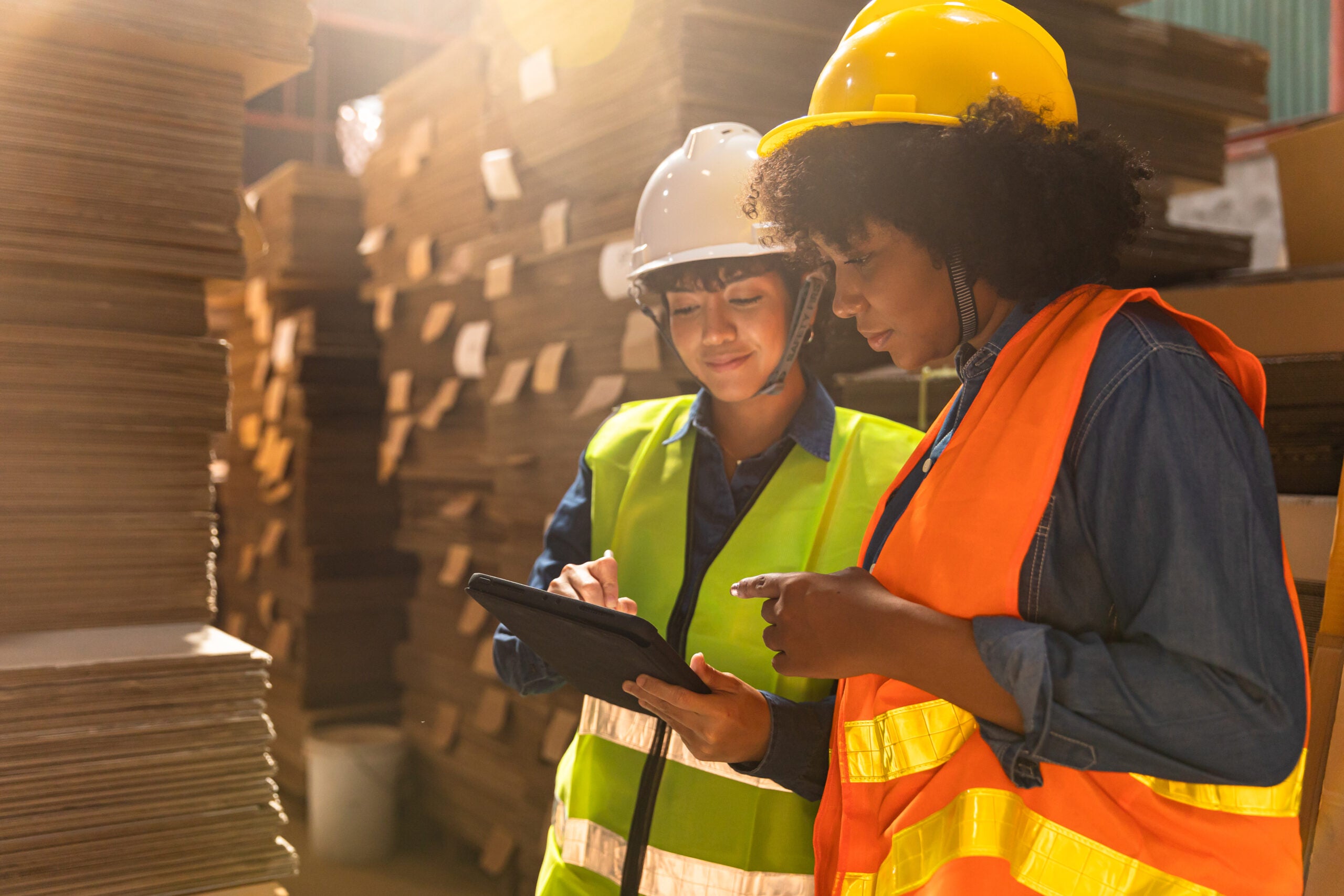 Engineer asian and african woman wearing safety helmet and vest holding clipboard and take note on the paper in the automotive part warehouse.Products and corrugated cardboard. Factory for the manufacture and processing of paper.