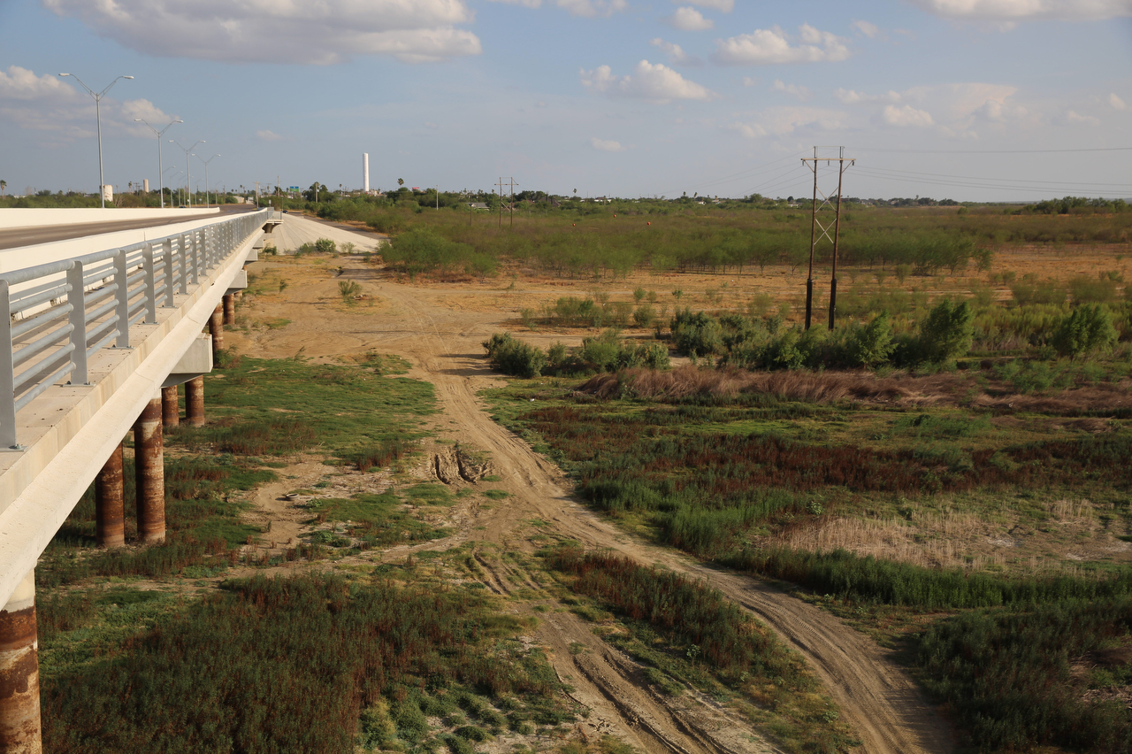 A bridge crosses a dry, scrub-covered creek bed without a hint of water in sight, under a big partly cloudy sky.