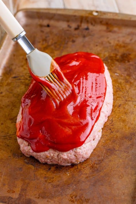 Ketchup being spread on a raw meatloaf.