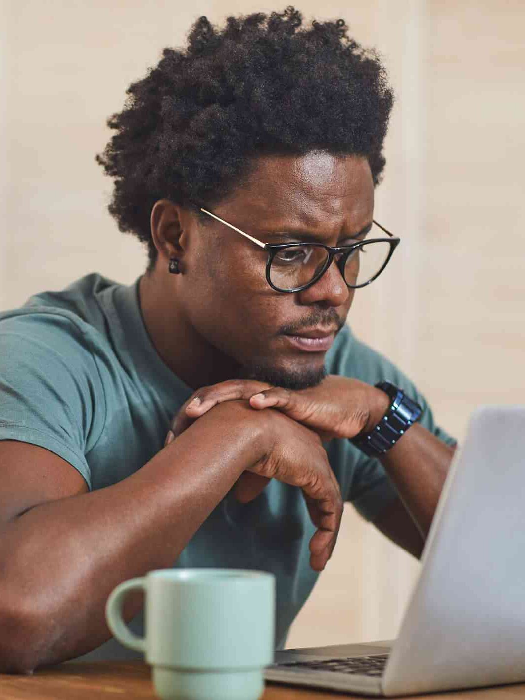 A person with glasses sits at a table, looking at a laptop screen, with a green mug nearby.