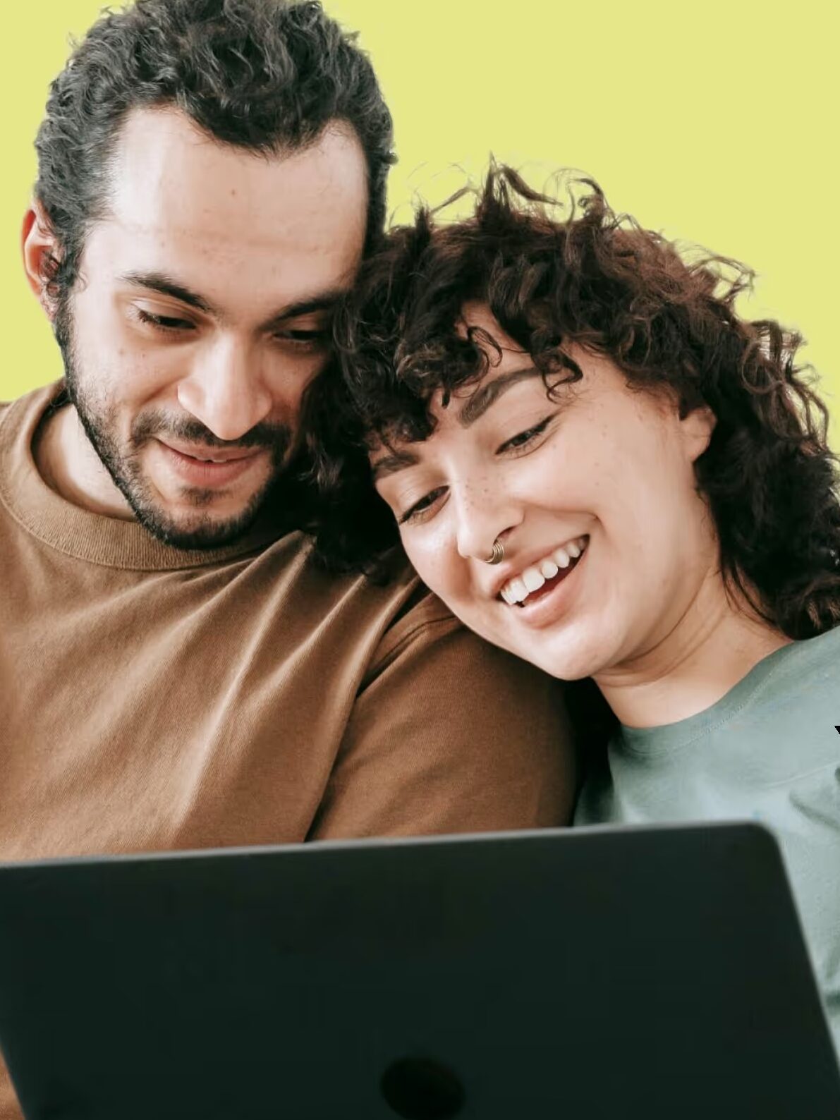 A man and woman sit closely together, smiling while looking at a laptop screen, against a yellow background.