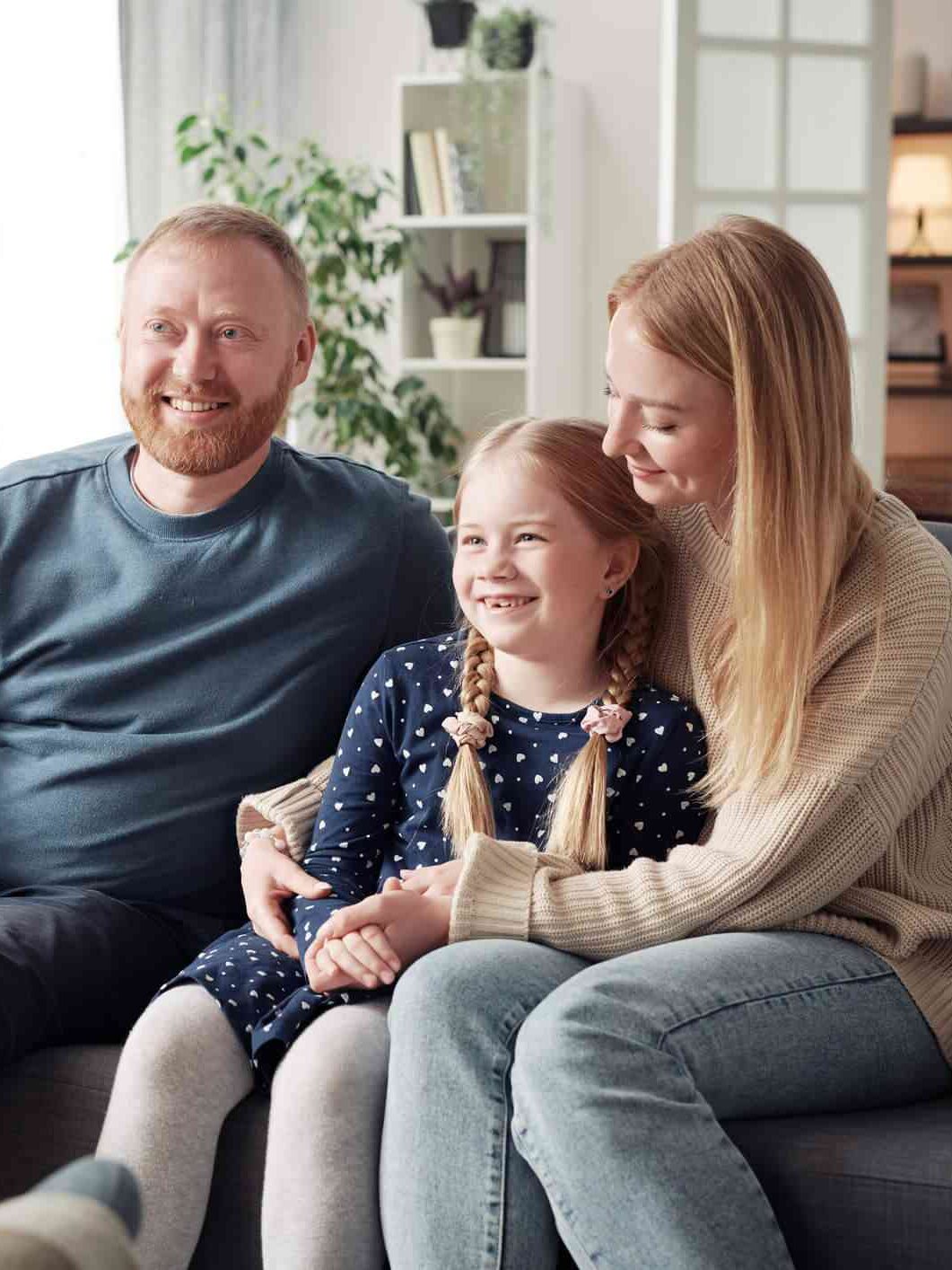 A family of three sits on a sofa during a counseling session. The man and woman smile while the young girl, with braided hair, sits between them. Another person is partially visible taking notes.