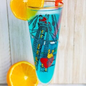 A blue AMF cocktail drink garnished with an orange slice that's in a vintage pirates motif glass