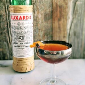 Martinez Cocktail in a silver coupe glass with a bottle of Luxardo behind it