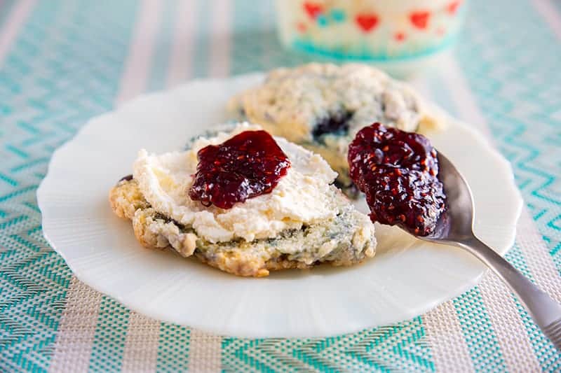 white plate with clotted cream on a scone topped with some jam, underneath is a pink and blue printed table cloth, clotted cream in a heart patterned Pyrex container in background