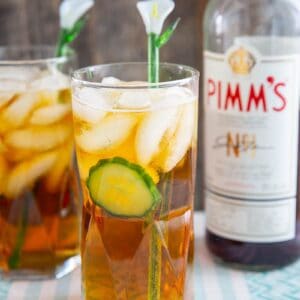 Pimm's cup cocktail in a tall glass with cucumber, and a glass lily flower stir stick.Bottle of Pimm's in background.