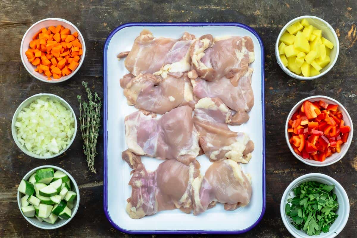 ingredients for chicken stew including boneless skinless chicken thighs, onion, garlic, carrots, red bell peppers, zucchini, potato, parsley, and fresh thyme.