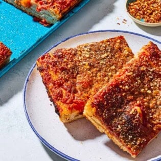 A close up of 2 slices of sfincione sicilian pizza on a plate. Next to this is the rest of the pizza on a sheet pan and a small bowl of red pepper flakes.