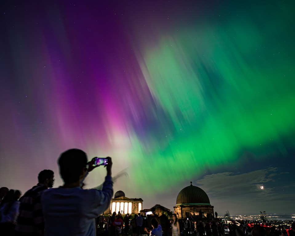 A crowd of people on Calton Hill in Edinburgh, Scotland, watching the aurora borealis.  A person photographs the aurora with a smartphone.