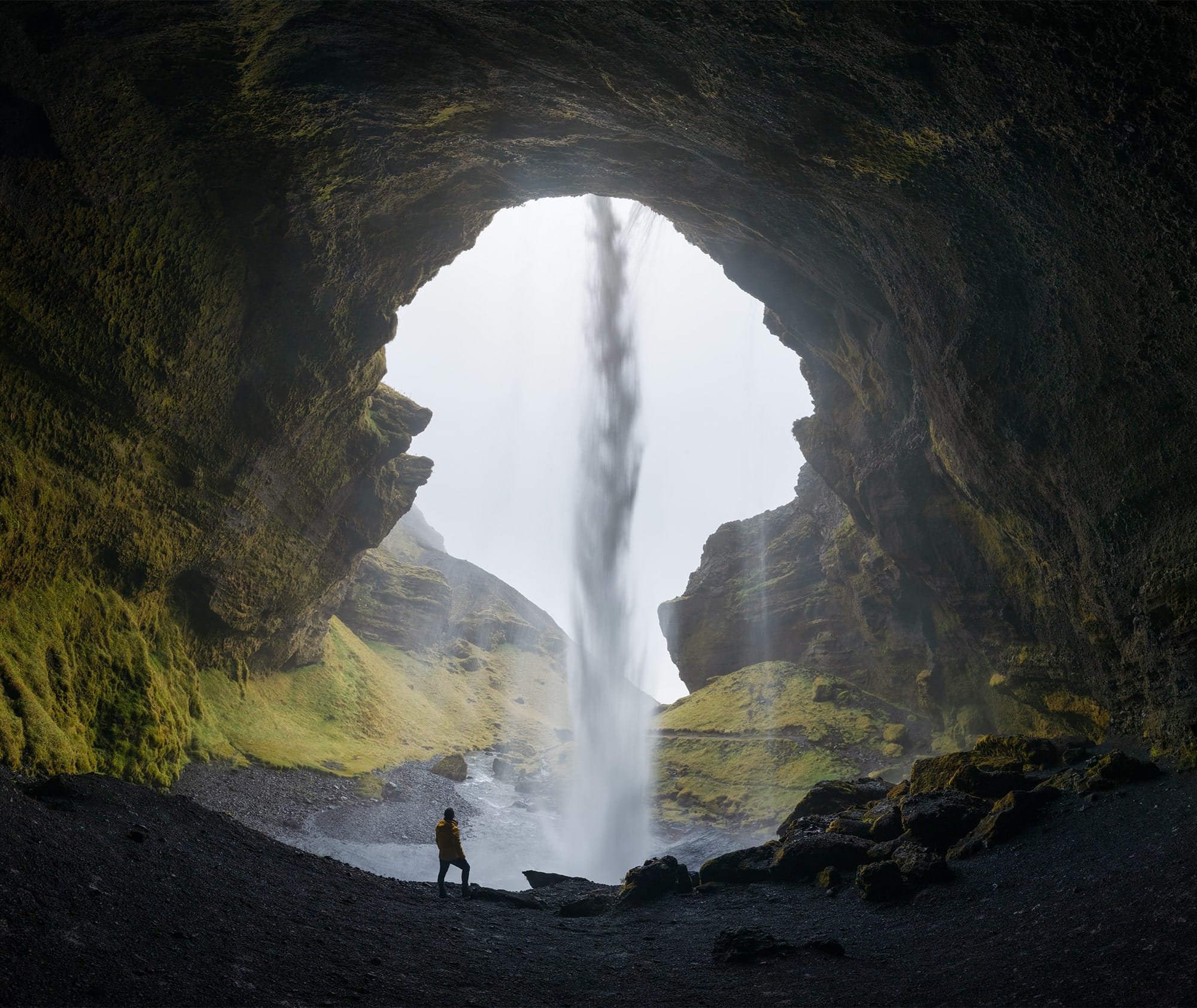 a figure stands in the opening of a cave, its silhouette accentuated against a waterfall