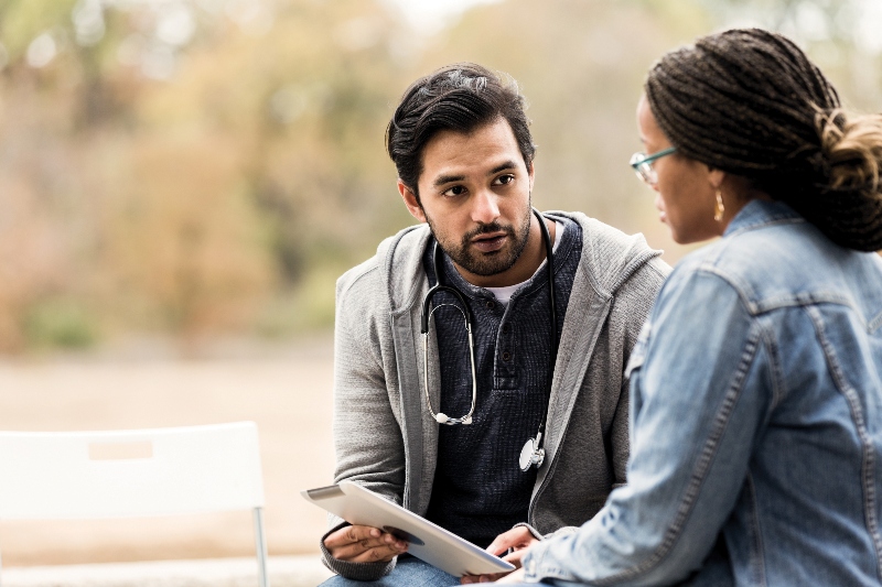 A male doctor with a stethoscope sits with a female patient in a denim jacket.