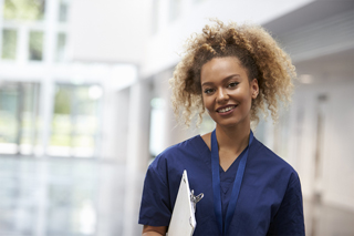 Nurse standing in a hospital corridor, smiling at the camera, holding a clipboard.