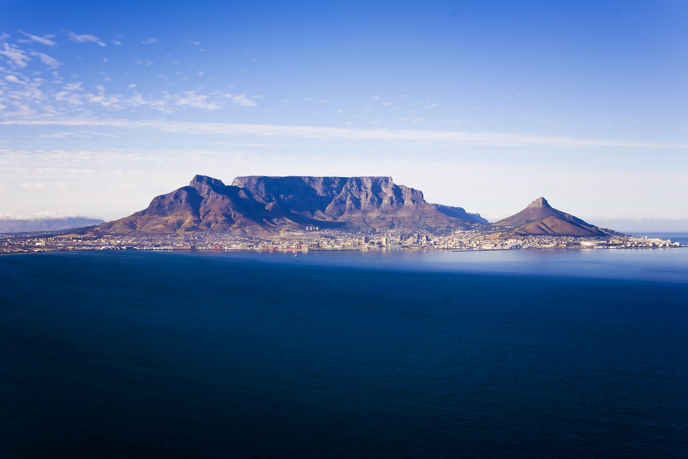 A panoramic view of Cape Town with Table Mountain and Lion's Head in the background. The cityscape includes buildings and roads at the base of the mountains, with the ocean in the foreground. The clear blue sky and expansive view of the ocean highlight the beauty of this iconic landscape. Visiting Table Mountain is one of the cheap things to do in Cape Town.