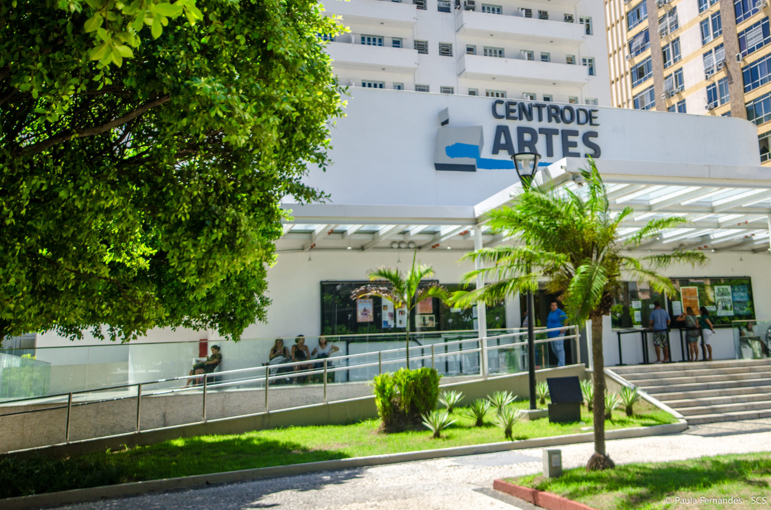 Foto da fachada do Centro de Artes da UFF. Localizado no térreo da Reitoria, na fachada frontal está o logotipo onde se lê 'Centro de Artes', com escadas e rampa para acesso. A frente desta, árvores do jardim da reitoria.