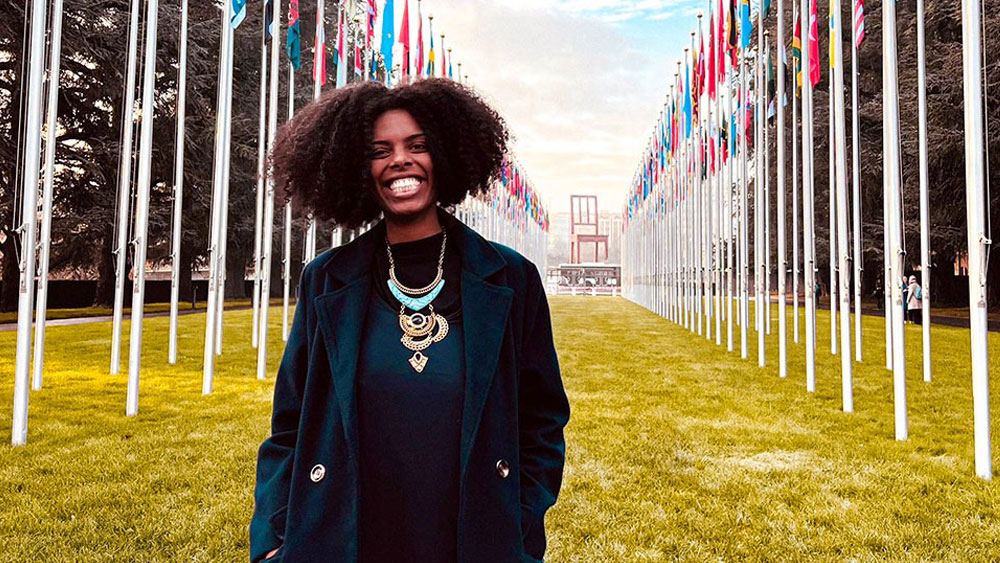 Monique stands in an open space with 2 rows of flags behind her