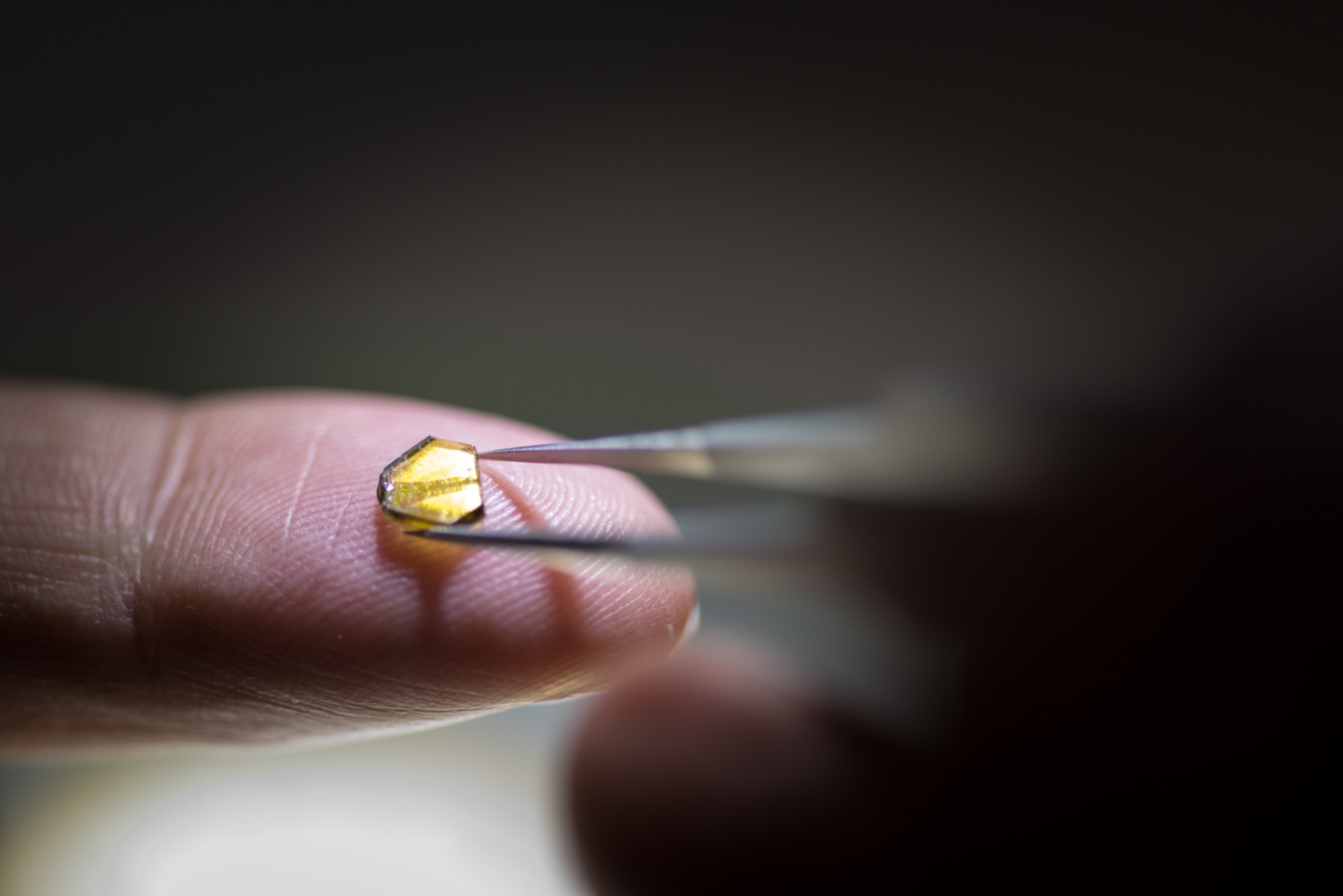 Close-up of a small, yellow diamond structure held with tweezers. The diamond lies on the tip of a finger. The background is blurred and dark, putting the diamond and the tweezers in focus.