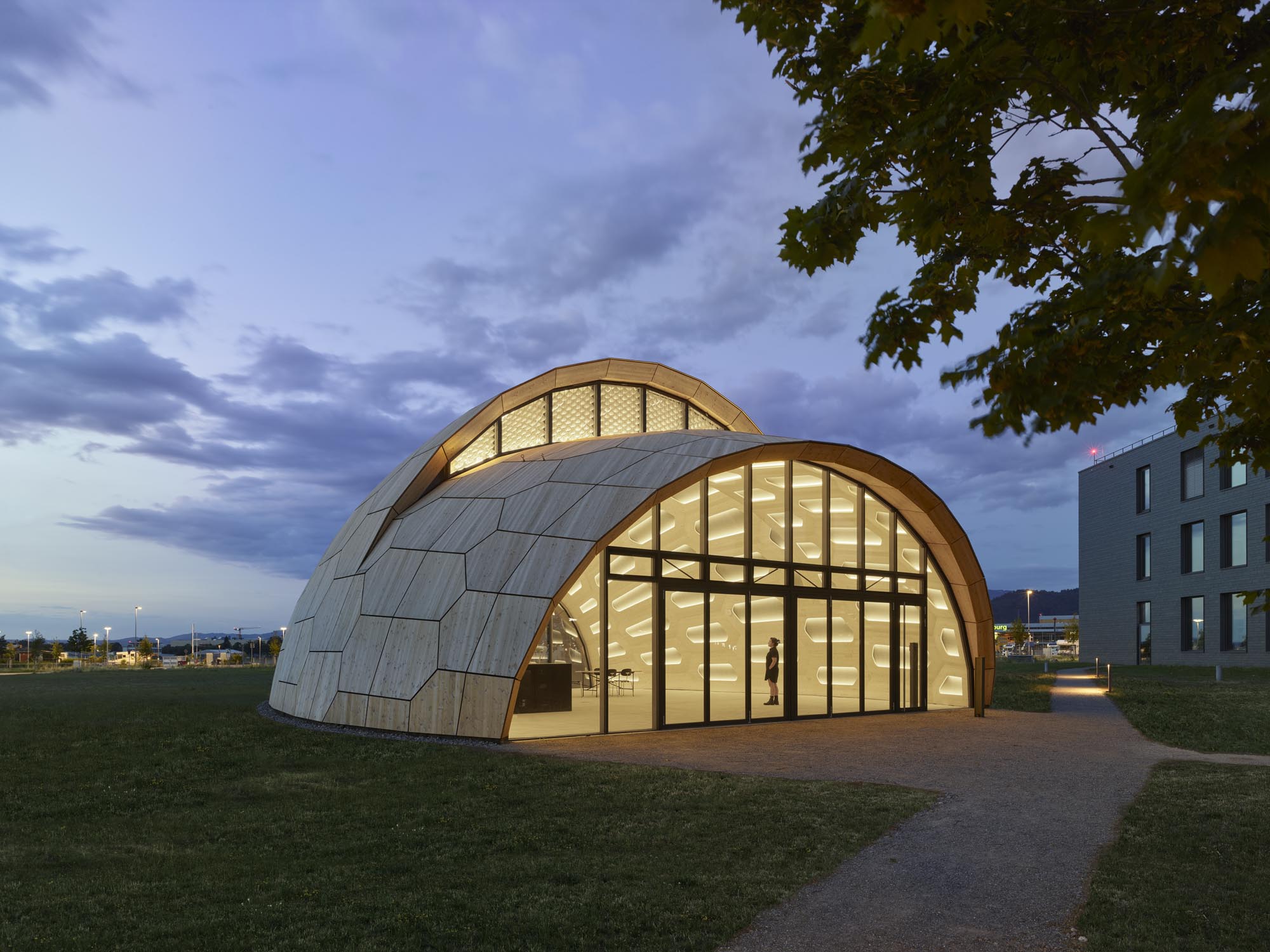 A modern, dome-shaped wooden structure with large glazed entrances, illuminated from the inside, standing in an evening landscape. In the background, a building and trees are visible, while the sky is covered with clouds.