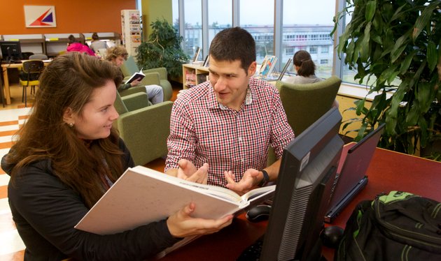 Femme avec cahier ouvert à la main et homme qui discutent à la bibliothèque.