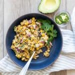 A blue bowl sitting on a striped towel with a corn salad in it and a spring of cilantro on the side.