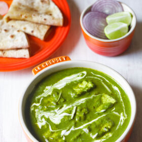 palak paneer topped with cream, garnished with ginger julienne and served in an orange bowl on a white board with roti kept on a dark orange plate on top left and a small orange bowl with round sliced onions and lemon wedges on top